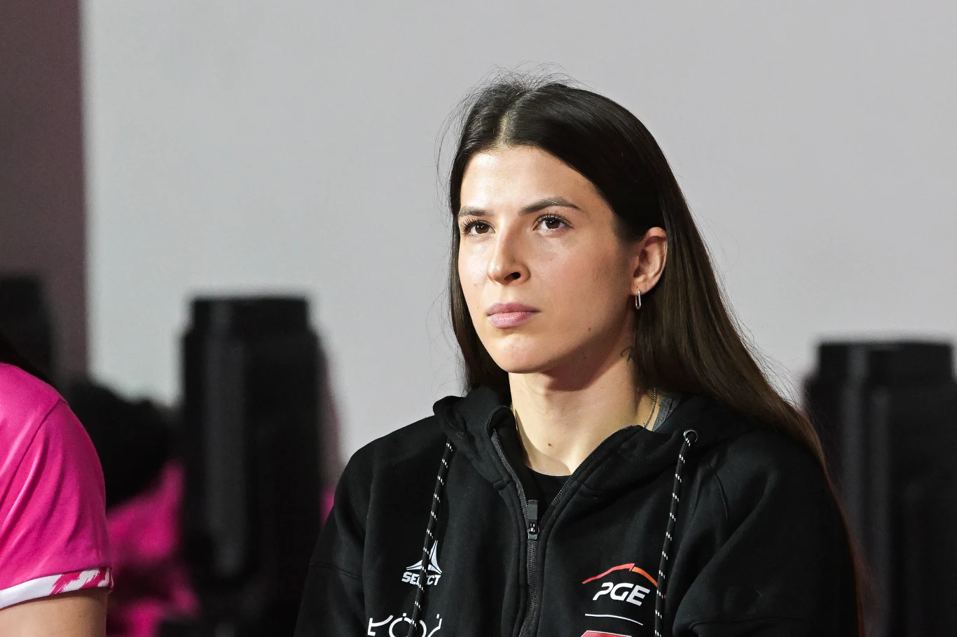 Młoda kobieta o długich ciemnych włosach, ubrana w czarną bluzę sportową z logo PGE i emblematami sponsora, siedzi na tle rozmazanego tła i patrzy przed siebie z poważnym wyrazem twarzy.