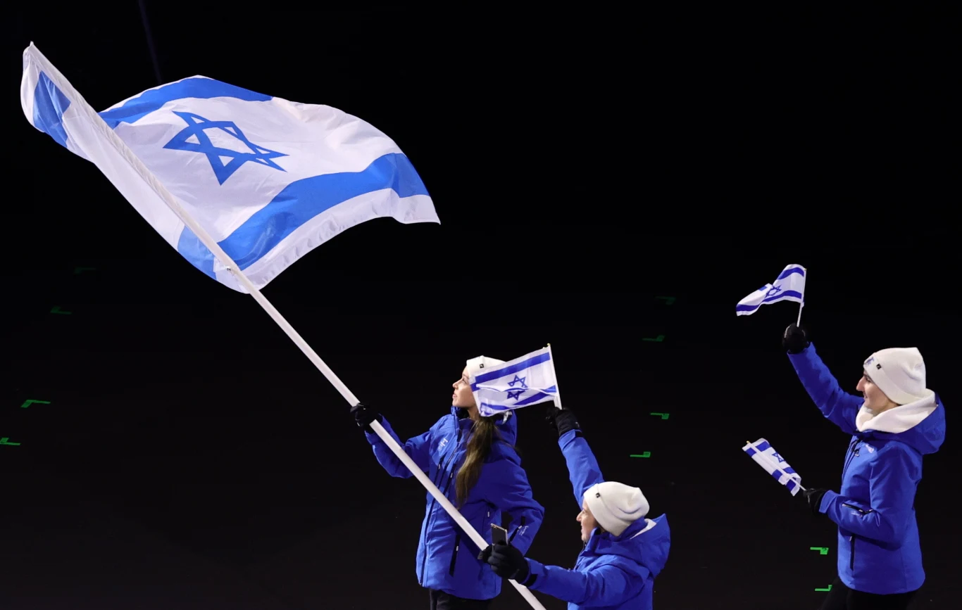 Trzy osoby w niebieskich kurtkach i białych czapkach trzymają izraelskie flagi na tle ciemnego, jednolitego tła. Największa flaga, trzymana przez osobę po lewej, przyciąga najwięcej uwagi. Wszyscy uczestnicy wyglądają na zaangażowanych i skoncentrowanych.
