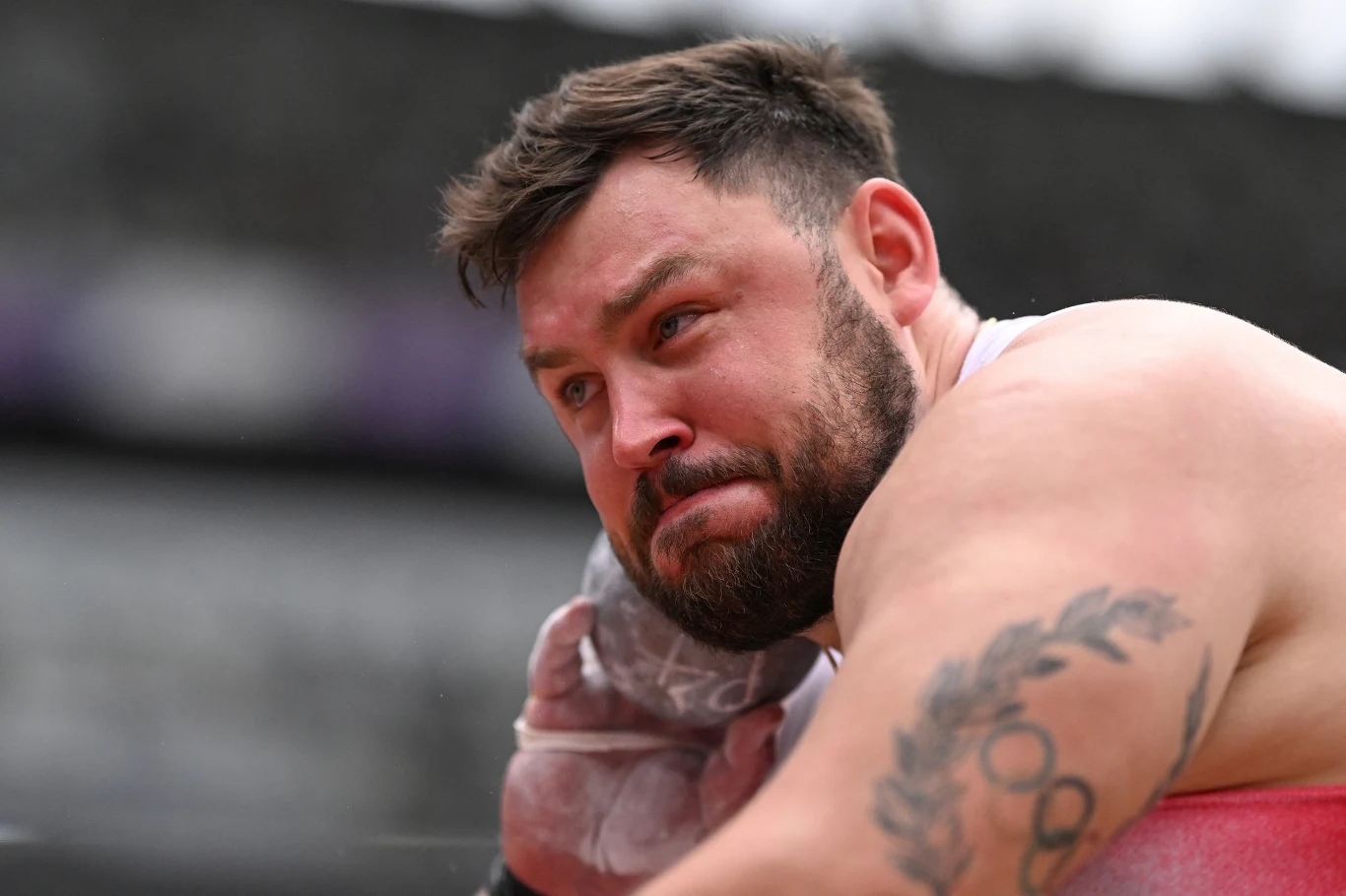 Sportowiec w trakcie wykonywania pchnięcia kulą, skupiona twarz, napięte mięśnie ramion i widoczny tatuaż na ręce.