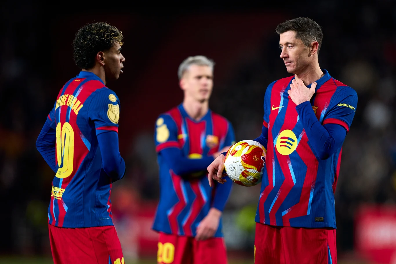 Lamine Yamal i Robert Lewandowski Dwóch piłkarzy drużyny FC Barcelona w charakterystycznych granatowo-bordowych strojach rozmawia ze sobą na boisku, jeden z nich trzyma piłkę w dłoniach, w tle kolejny zawodnik.