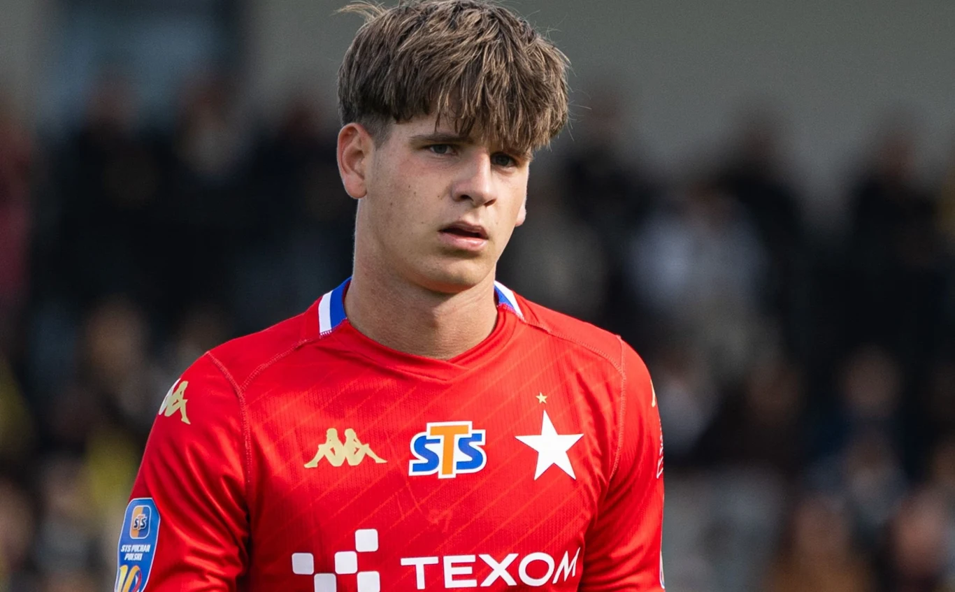 Szymon Kawała Młody piłkarz w czerwonym stroju sportowym klubu Wisła Kraków, z widocznymi logotypami sponsorów, stoi na tle rozmytego stadionu podczas meczu.