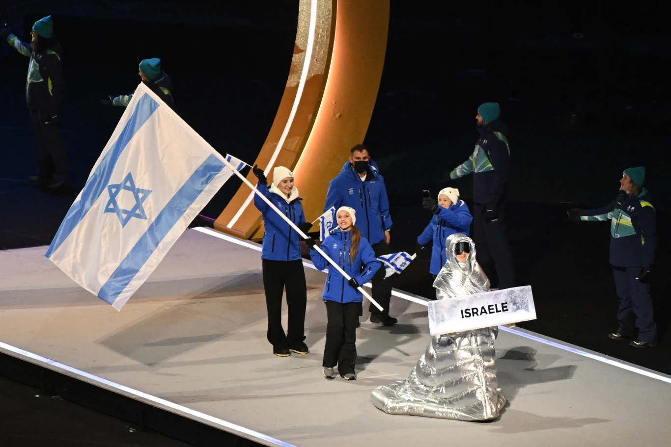Reprezentacja Izraela podczas ceremonii otwarcia igrzysk, kilka osób ubranych w oficjalne stroje maszeruje z dużą flagą oraz tabliczką z nazwą kraju, wokół nich inni członkowie delegacji.