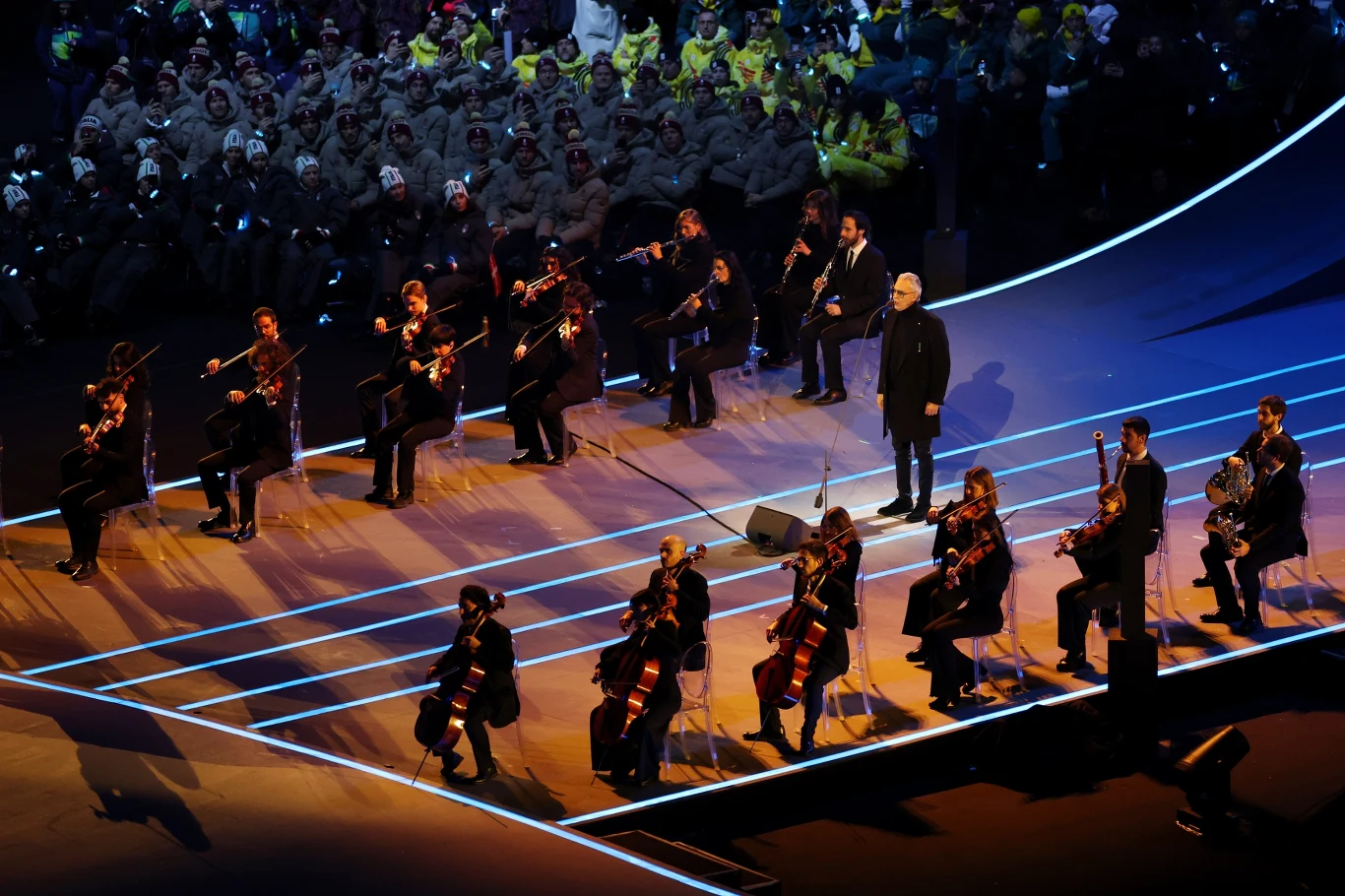 Andrea Bocelli na ceremonii otwarcia igrzysk olimpijskich Grupa muzyków grających na instrumentach smyczkowych i dyrygent występują na oświetlonej scenie, w tle widoczna jest publiczność ubrana w jednakowe stroje.