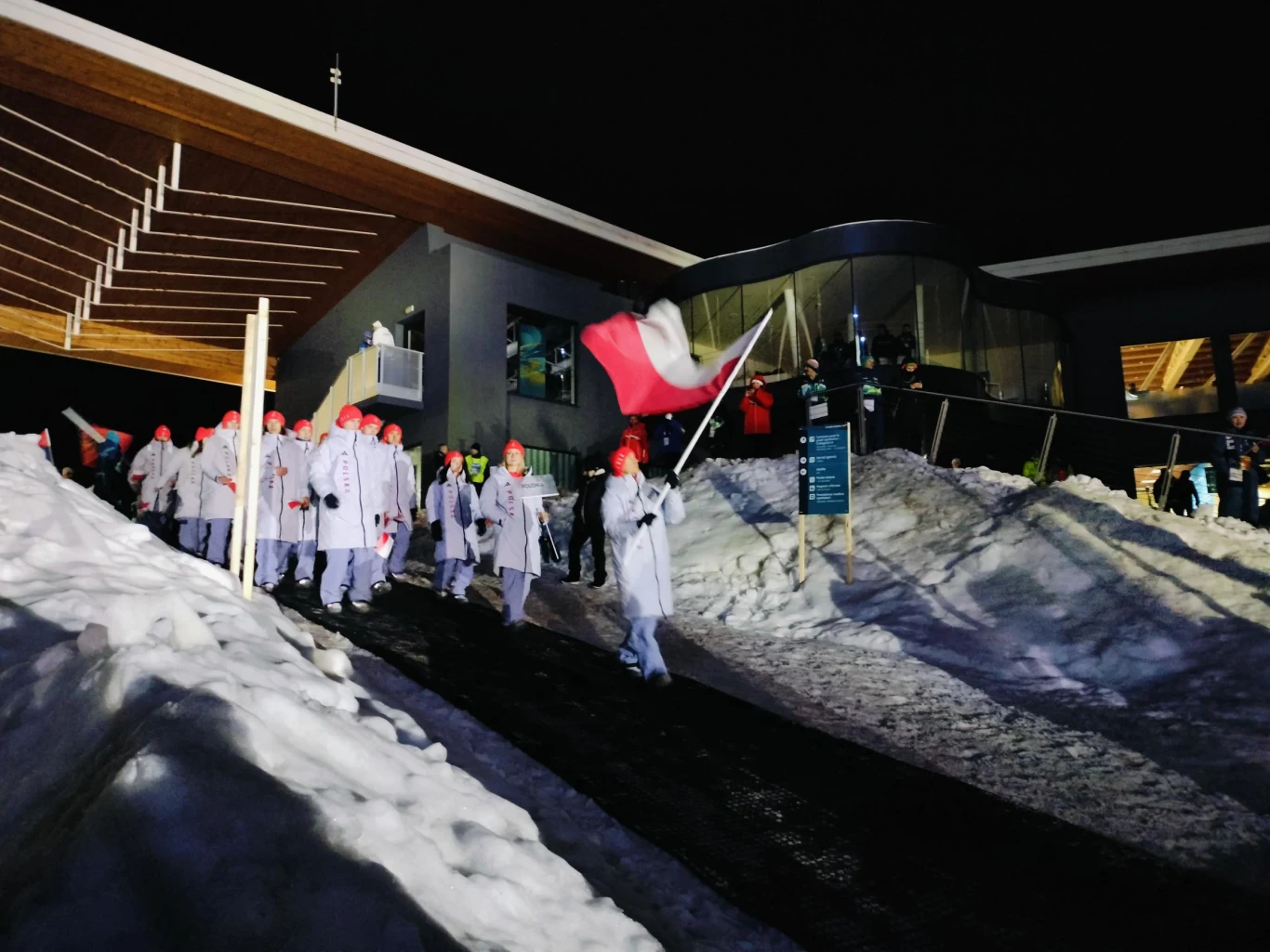 Grupa ludzi ubranych w białe kurtki i czerwone czapki maszeruje pośnieżonej alei, jedna osoba niesie biało-czerwoną flagę. Otoczenie stanowią nowoczesne budynki i nocne światło.