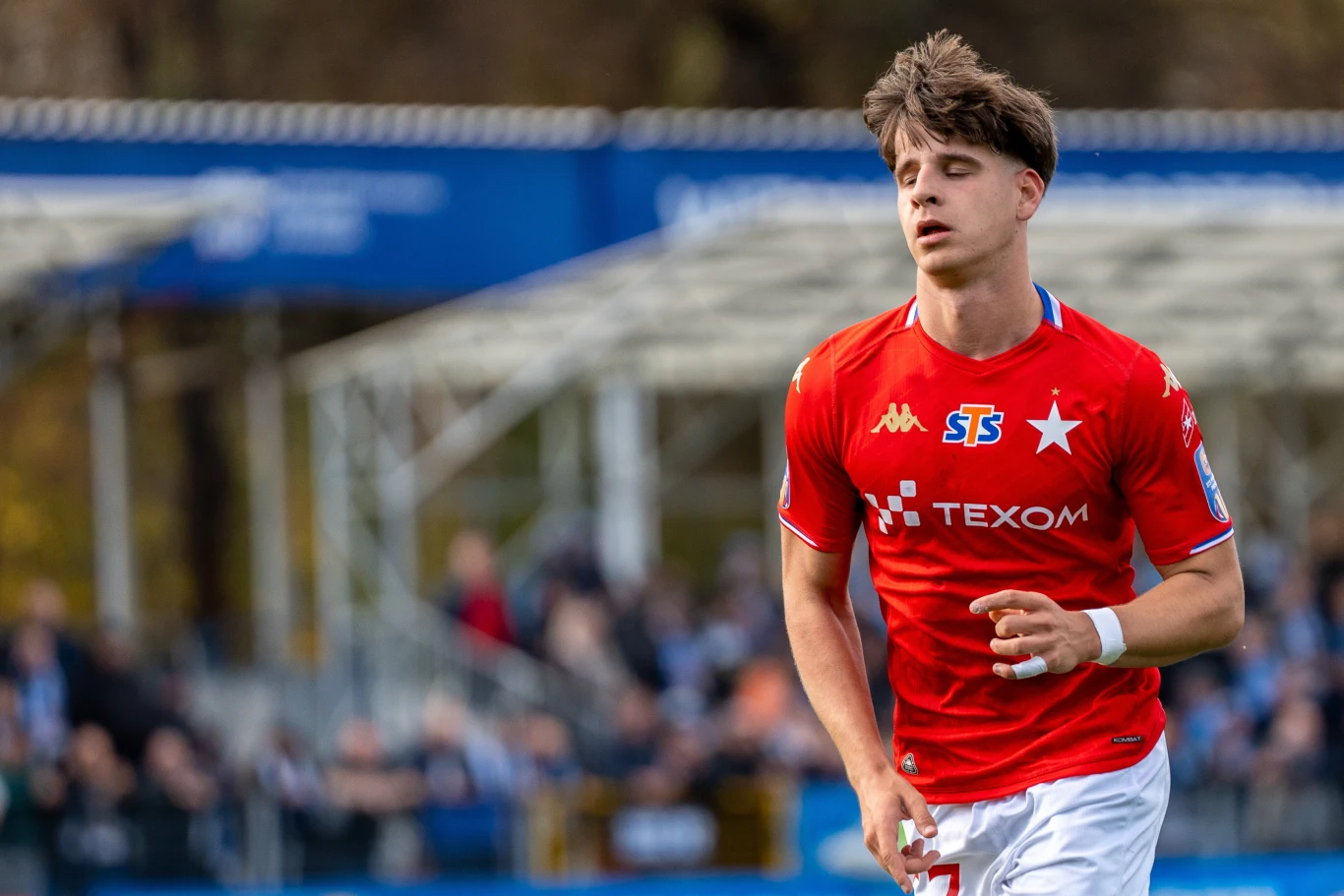 Szymon Kawala Młody piłkarz w czerwonej koszulce z białymi akcentami na tle trybun podczas meczu piłkarskiego, skupiony i w ruchu, z widocznymi sponsorami na stroju sportowym.