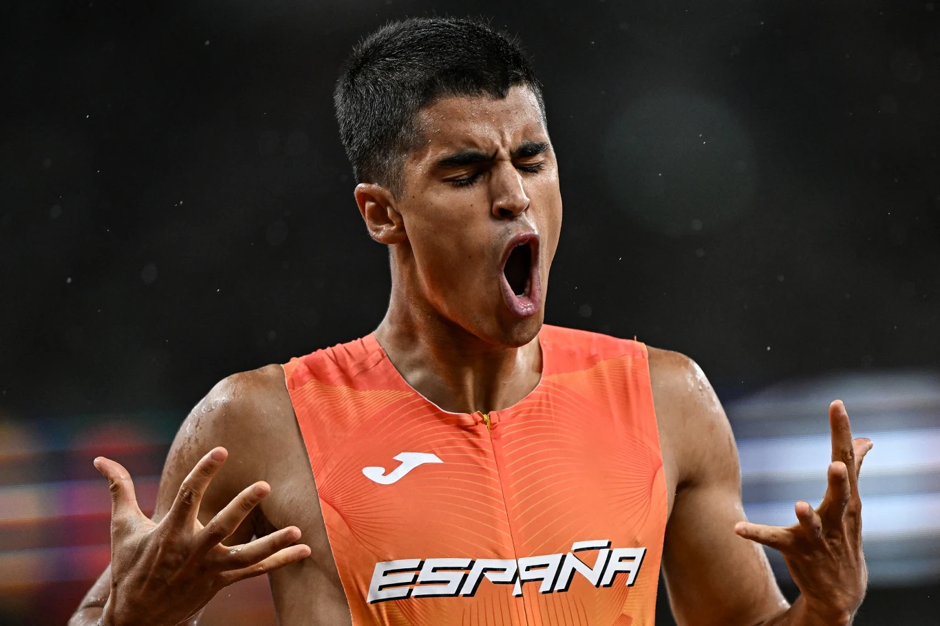 Młody lekkoatleta w stroju reprezentacji Hiszpanii ze ściśniętymi dłońmi i wyrazistą mimiką twarzy, ukazujący emocje związane z rywalizacją sportową.