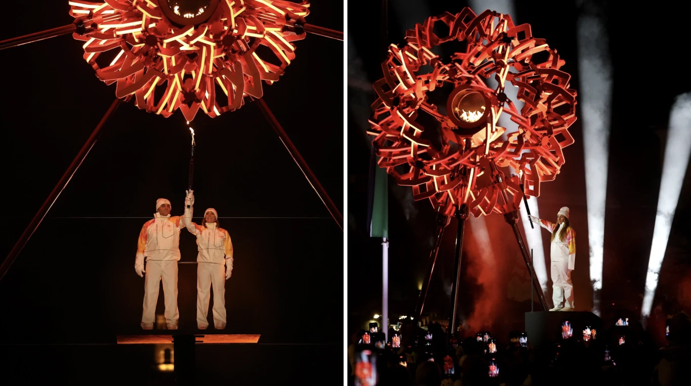 Zapłonęły znicze olimpijskie we Włoszech Dwoje sportowców w białych kombinezonach z pochodnią stoi pod oryginalną instalacją artystyczną w kształcie śnieżynki, otoczoną światłami sceny i liczną publicznością rejestrującą wydarzenie telefonami.