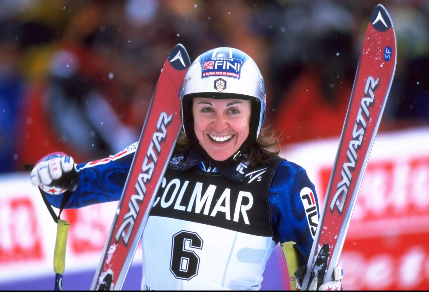 Deborah Compagnoni Uśmiechnięta narciarka w kombinezonie sportowym i kasku trzyma w rękach narty, na piersi widoczny numer startowy 6 i napis Colmar, w tle rozmazany tłum kibiców.