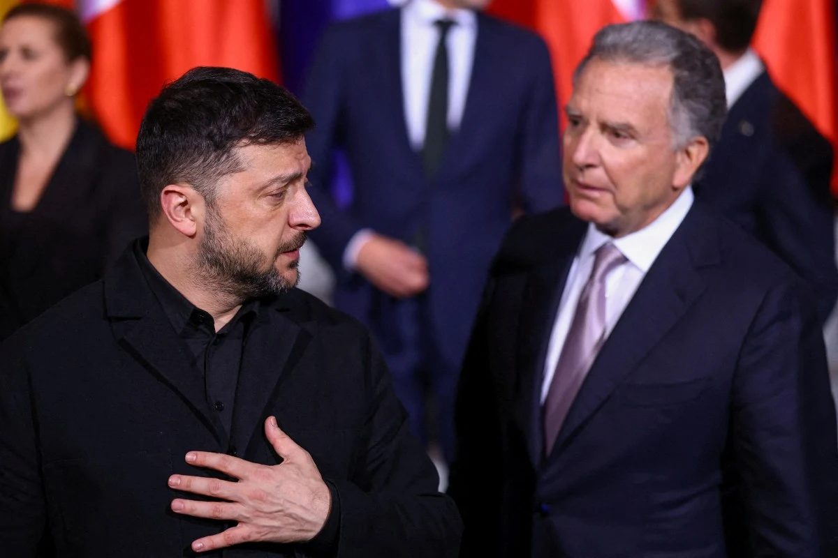 Dwóch mężczyzn w garniturach rozmawia ze sobą w oficjalnym otoczeniu, w tle widoczne flagi państwowe oraz inne osoby ubrane w eleganckie stroje.