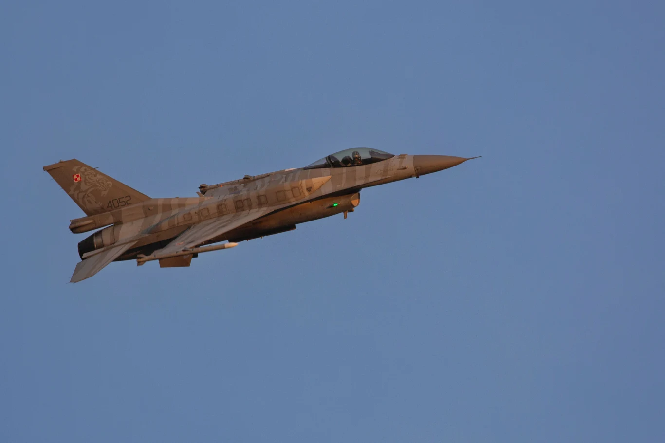 Myśliwiec wojskowy F-16 w locie na tle czystego, niebieskiego nieba, oznaczenia narodowe na ogonie samolotu.