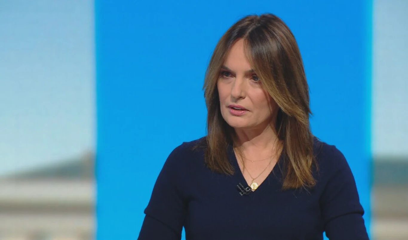 Kobieta o prostych, brązowych włosach w granatowej bluzce z mikrofonem przy kołnierzu stoi na tle niebiesko-białej ściany.