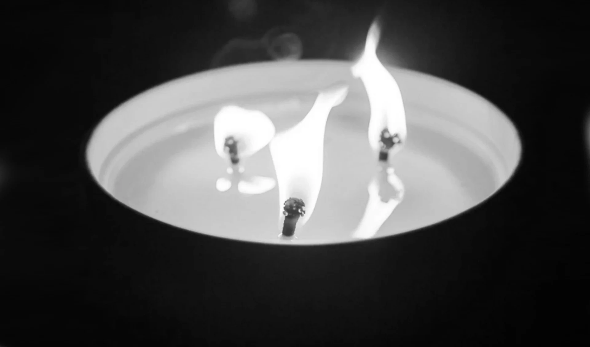 Płonąca świeca z trzema wyraźnie widocznymi kniotami, otoczona ciemnym tłem, ogień tworzy delikatne refleksy na powierzchni wosku.