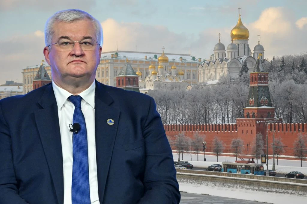 Mężczyzna w garniturze z krawatem na tle zimowego krajobrazu Kremla w Moskwie, na pierwszym planie widoczny fragment czerwonego muru i kilka przejeżdżających samochodów, w głębi złote kopuły soboru.