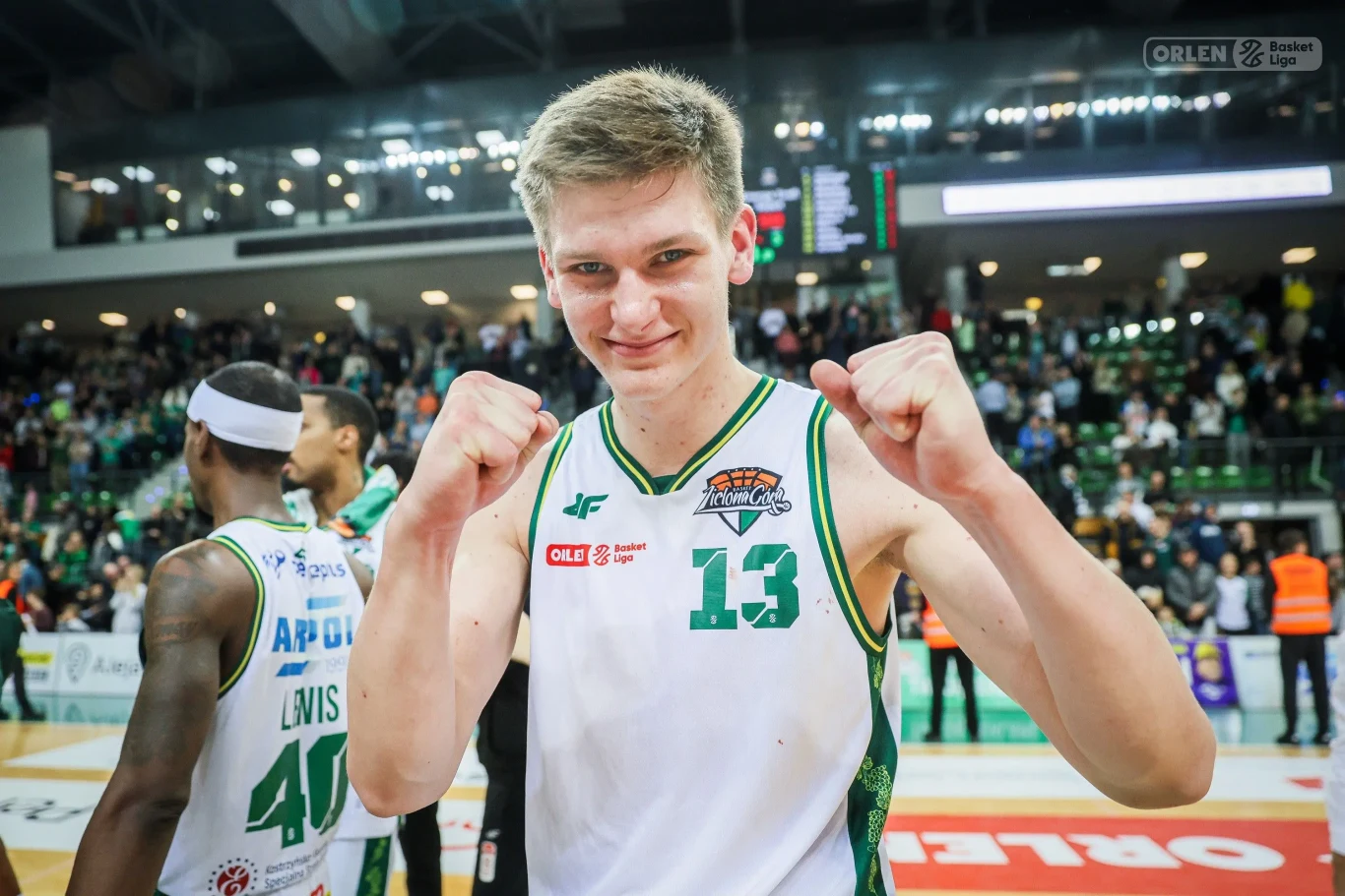 Sportowiec w białozielonym stroju drużyny koszykarskiej cieszy się po meczu, zaciśnięte pięści uniesione w triumfującym geście. W tle widać halę sportową, kibiców oraz innych zawodników.