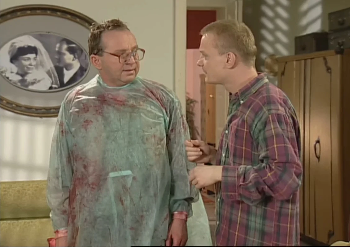 Dwaj mężczyźni w pomieszczeniu, jeden ubrany w poplamiony fartuch ochronny, drugi rozmawia z nim w swobodnej pozie. W tle widoczne zdjęcie ślubne w ramce na ścianie oraz szafa.