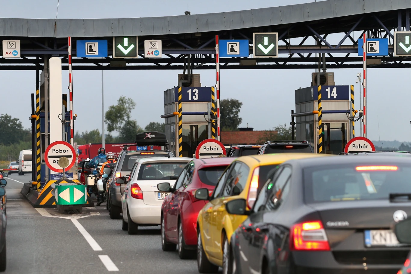 Samochody stojące w kolejce do bramek autostradowych podczas poboru opłat, widoczne oznakowanie, kilku motocyklistów oraz oznaczenia pasów i sygnalizacja świetlna.