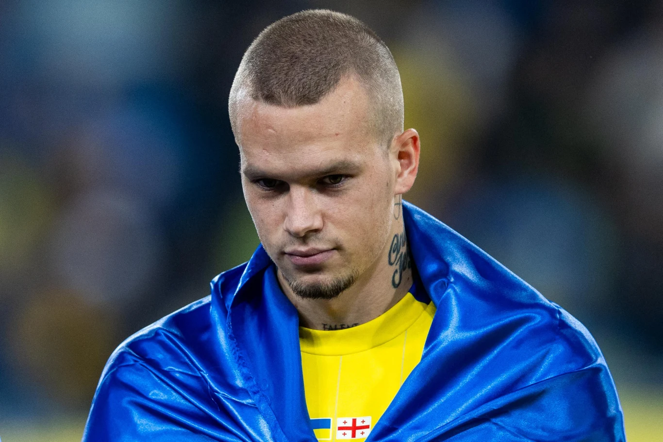 Młody mężczyzna z poważnym wyrazem twarzy, owinięty niebieską flagą ze żółtym elementem, w tle rozmazane światło stadionu.