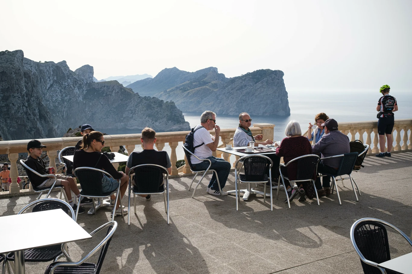 Branża gastronomiczna na Balearach odnotowała spadek liczby klientów w 2025 roku Grupa osób siedzi przy kawiarnianych stolikach na tarasie widokowym nad morzem, w tle wysokie, skaliste góry i rozległa tafla wody, jedna osoba stoi przy barierce.