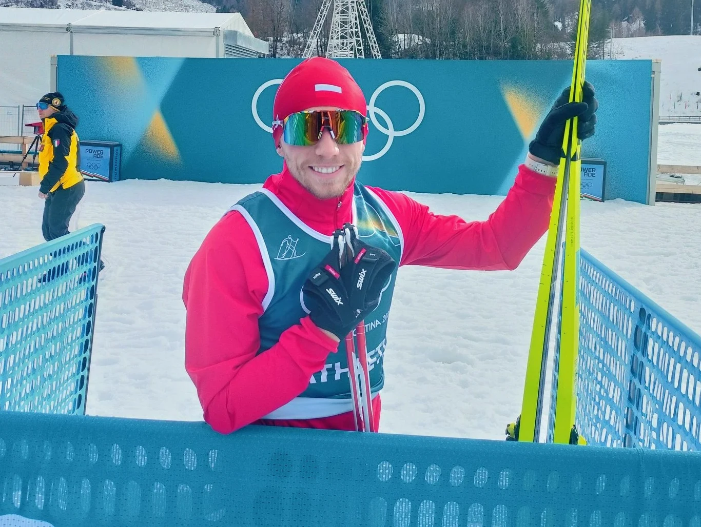 Sportowiec w czerwonym stroju narciarskim i okularach przeciwsłonecznych trzyma narty, stojąc na śniegu przy niebieskim ogrodzeniu. W tle widoczne logo olimpijskie oraz inna osoba w żółtym ubraniu.