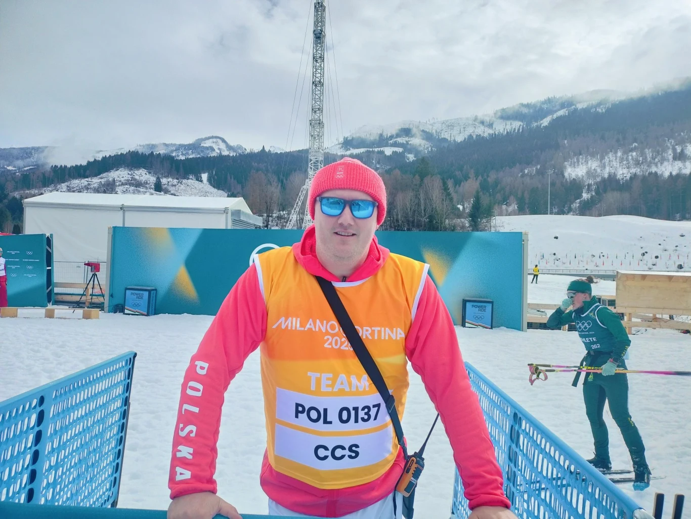 Członek polskiej drużyny w zimowym stroju sportowym i czerwonej czapce z niebieskimi okularami przeciwsłonecznymi stoi przy barierce na tle zaśnieżonych gór i infrastruktury sportowej. W tle widoczny inny narciarz na nartach biegowych.