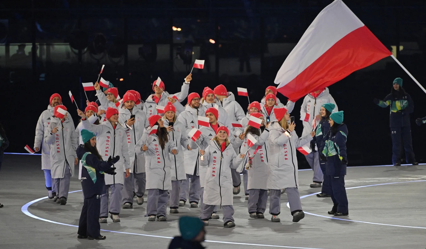Polska reprezentacja sportowa maszeruje podczas ceremonii otwarcia, ubrana w białe płaszcze i czerwone czapki, część osób macha biało-czerwonymi flagami, a jedna osoba niesie dużą polską flagę na czele grupy.