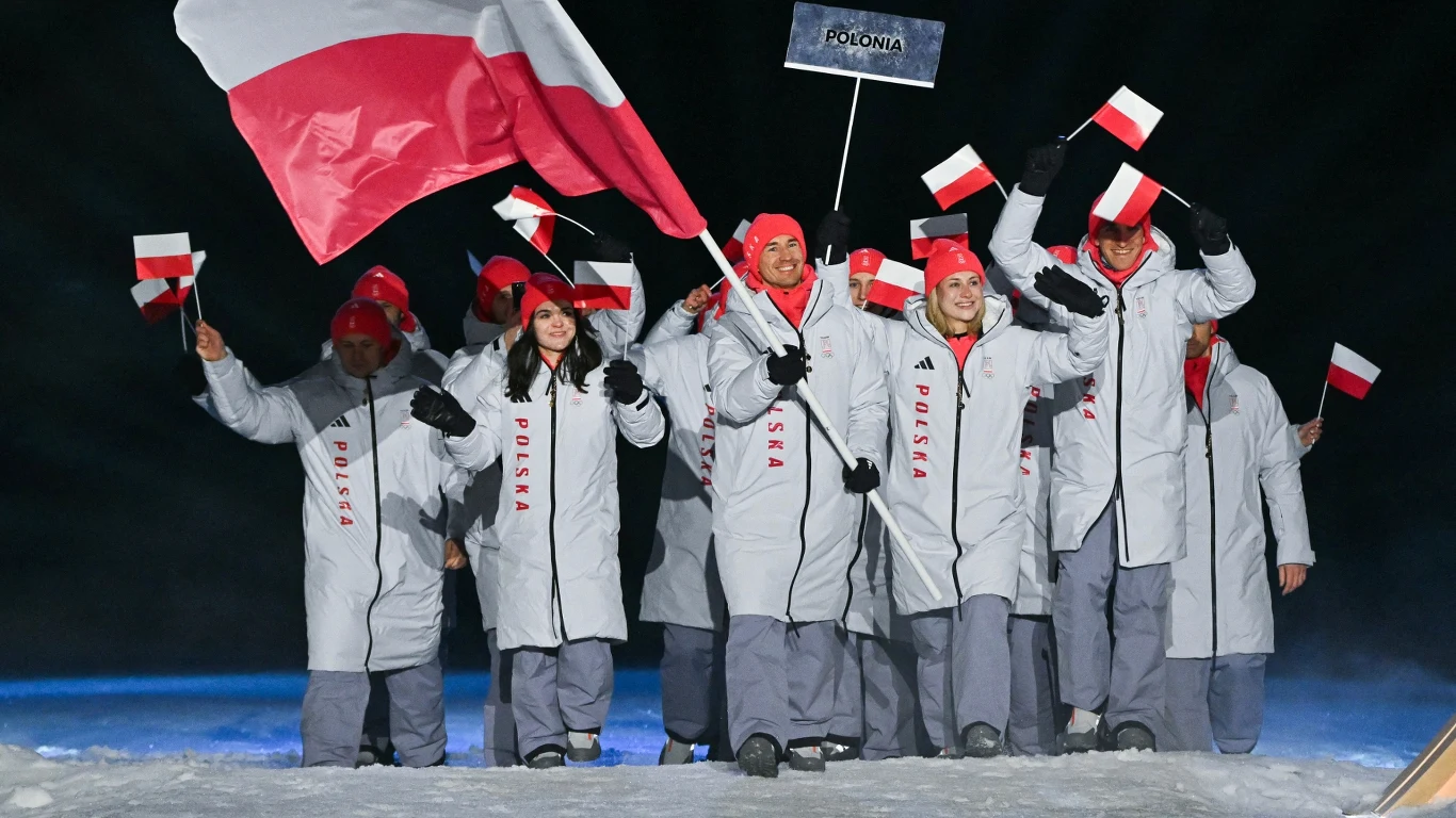 Grupa sportowców ubranych w białe kurtki i czerwone czapki trzyma małe flagi Polski oraz dużą biało-czerwoną flagę, idąc wspólnie po śniegu w jasno oświetlonym miejscu, prowadząc tabliczkę z napisem Polska.