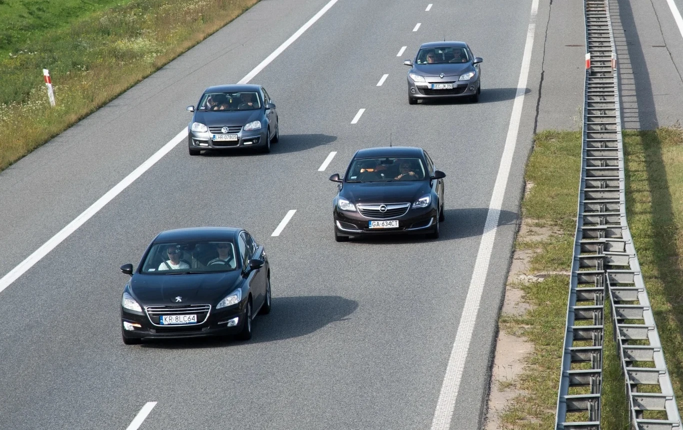 Czy wyprzedzanie prawym pasem na autostradzie jest dozwolone? Pięć samochodów osobowych jadących po czteropasmowej autostradzie z barierką ochronną po prawej stronie, jasna i sucha nawierzchnia, otoczenie zieleni oraz pas awaryjny.