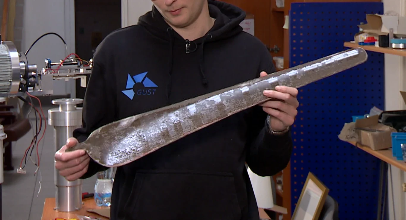 Osoba trzyma w rękach wydłużony, metaliczny element o kształcie łopaty lub płata, znajdując się w warsztacie lub laboratorium technicznym. W tle widoczne narzędzia oraz części elektroniczne.