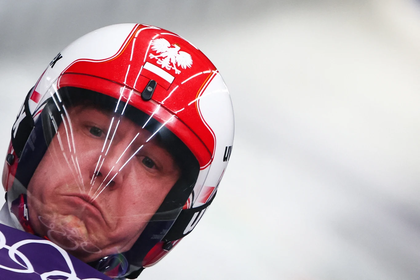 Zbliżenie na sportowca w kasku z orłem białym i flagą Polski, jadącego na sankach podczas zawodów zimowych, skupiona i napięta twarz widoczna przez szybkę kasku.