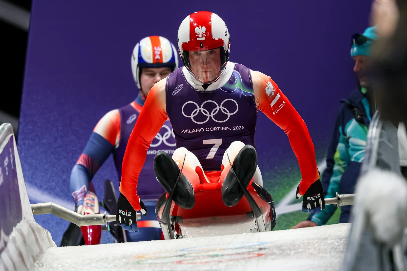 Zawodnik w stroju sportowym i czerwonym kasku, z numerem startowym, podczas startu na torze saneczkarskim podczas Zimowych Igrzysk Olimpijskich; w tle drugi sportowiec i oficjalne logo igrzysk.