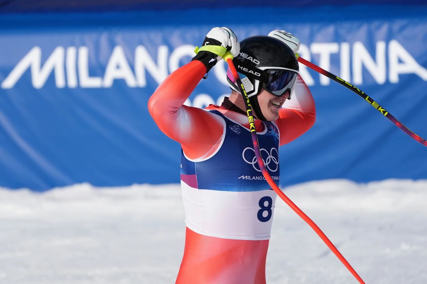 Narciarz alpejski w kombinezonie startowym i goglach, unoszący kijki do góry, tło z banerem zawodów olimpijskich Milano Cortina.