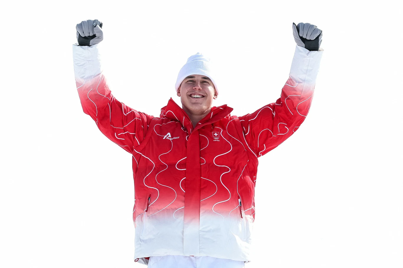 Uśmiechnięty sportowiec w czerwono-białej kurtce oraz czapce i rękawiczkach unosi obie ręce w geście zwycięstwa, wyraźna radość i duma na tle jasnego, zimowego krajobrazu.