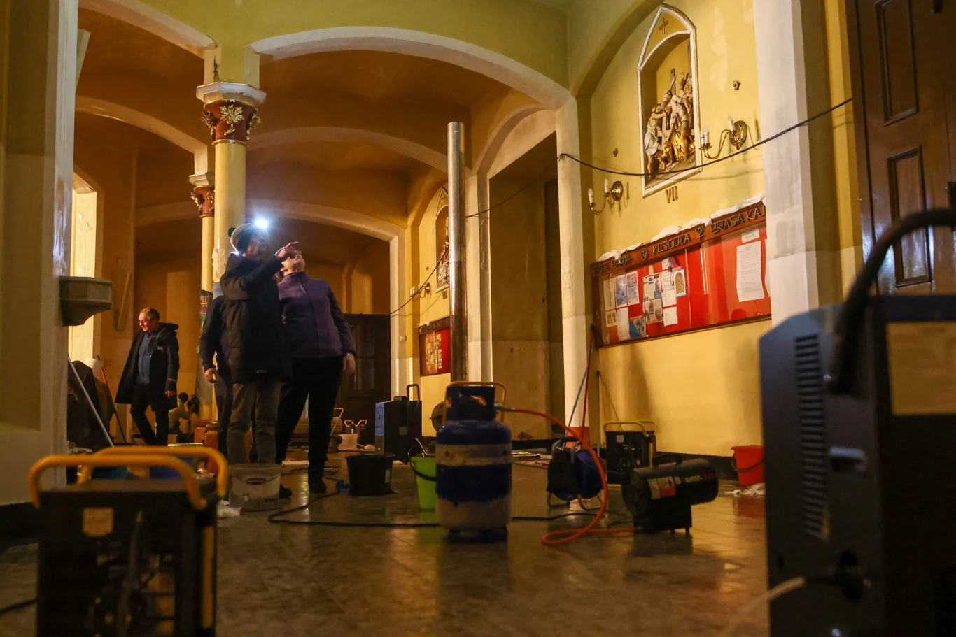 Wnętrze budynku o żółtych ścianach z wysokimi łukami, na podłodze rozstawiony sprzęt techniczny, kilka osób ubranych w kurtki i latarki czołowe ogląda rozmieszczone urządzenia. Na ścianie widoczna tablica informacyjna oraz wnęka z rzeźbą religijną.