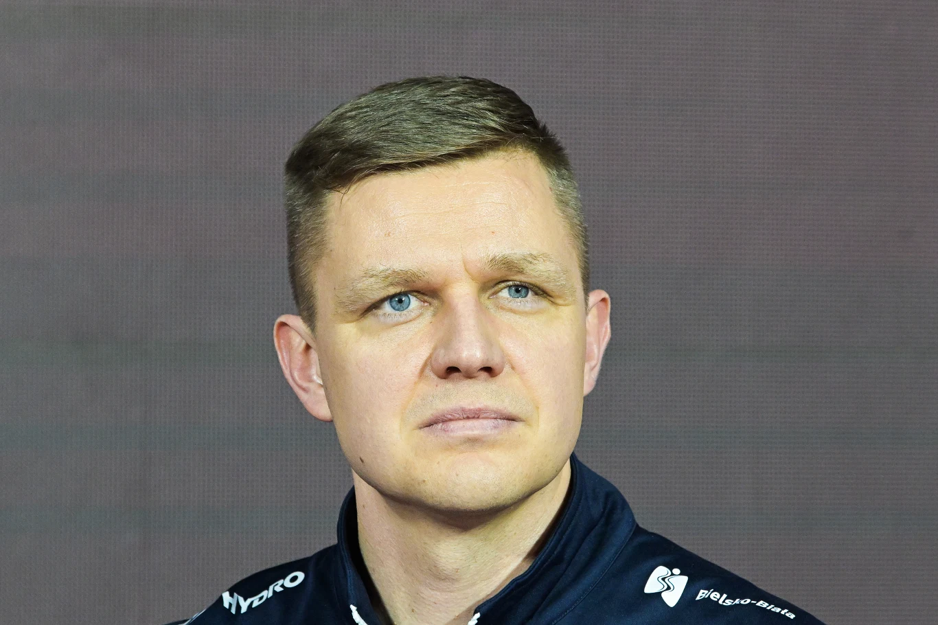Mężczyzna o krótkich, jasnych włosach i niebieskich oczach ubrany w granatową kurtkę sportową na tle jednolitego, ciemnoszarego tła. Wyraz twarzy poważny, skierowany lekko w bok.