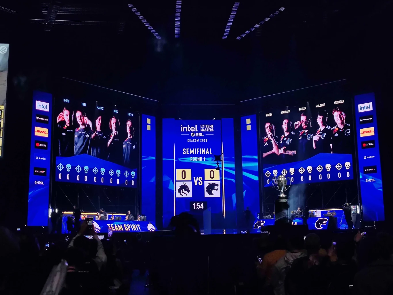 Duża scena e-sportowa z ogromnymi ekranami prezentującymi dwa zespoły rywalizujące w półfinale turnieju. Po bokach widoczne są drużyny siedzące przy komputerach oraz liczba punktów obu zespołów, na środku zegar odlicza czas do rozpoczęcia meczu.