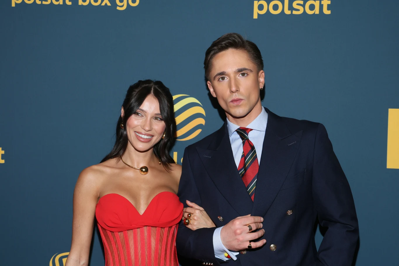 Natsu i Wojciech Kucina Para elegancko ubranych osób pozuje razem na tle ścianki z logo Polsat Box Go, kobieta w czerwonym gorsecie i mężczyzna w granatowym garniturze z krawatem w paski, oboje uśmiechnięci.