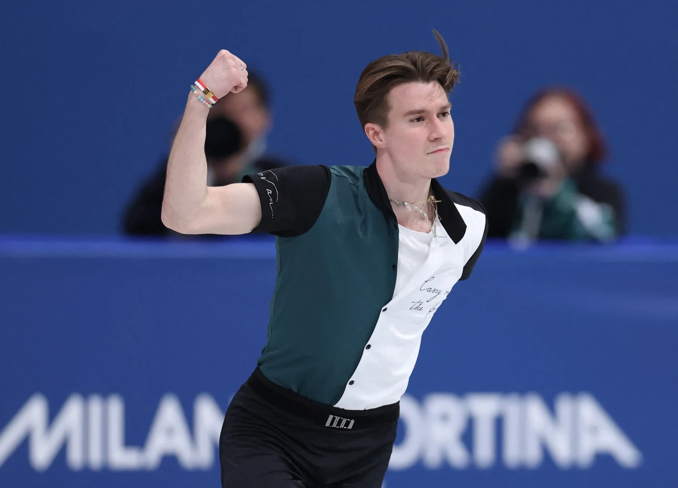 Łyżwiarz figurowy w dynamicznej pozie na lodowisku, z determinacją na twarzy, w tle rozmyta publiczność.