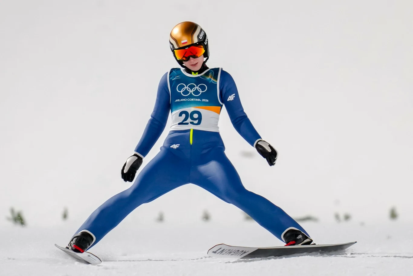 Anna Twardosz Skoczek narciarski w niebieskim stroju z numerem startowym 29 oraz złotym kaskiem ląduje na śniegu podczas zawodów olimpijskich, rozstawiając szeroko narty.
