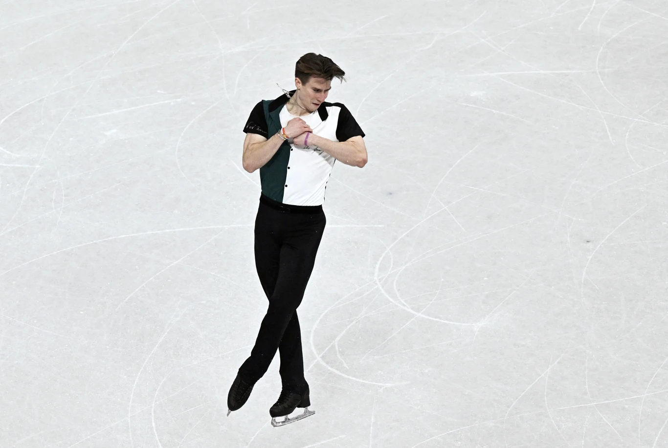 Łyżwiarz figurowy w eleganckim stroju wykonujący skomplikowany element na lodowisku, otoczony śladami po łyżwach na jasnej tafli.