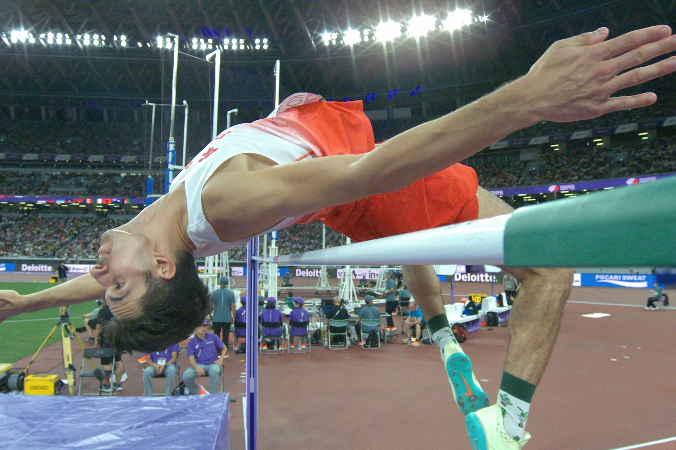 Lekkoatleta w trakcie skoku wzwyż nad poprzeczką na stadionie lekkoatletycznym, publiczność w tle oraz jasno oświetlona arena sportowa.