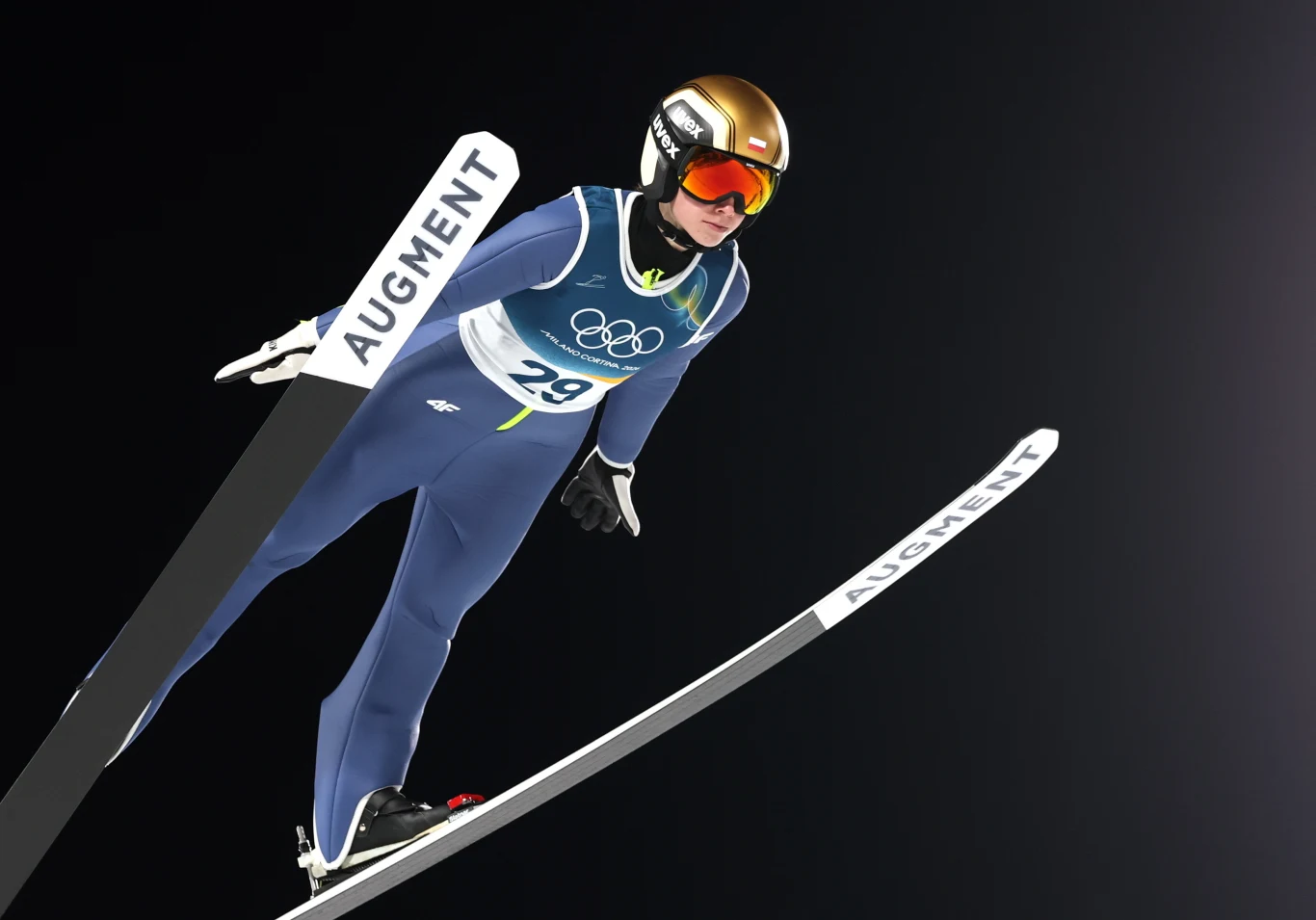 Zawodnik w stroju sportowym podczas skoku narciarskiego, ubrany w niebieski kombinezon, złoty kask i gogle, na tle ciemnego nieba, z widocznymi nartami z napisem AUgment oraz numerem startowym 29 i olimpijskimi symbolami na kamizelce.