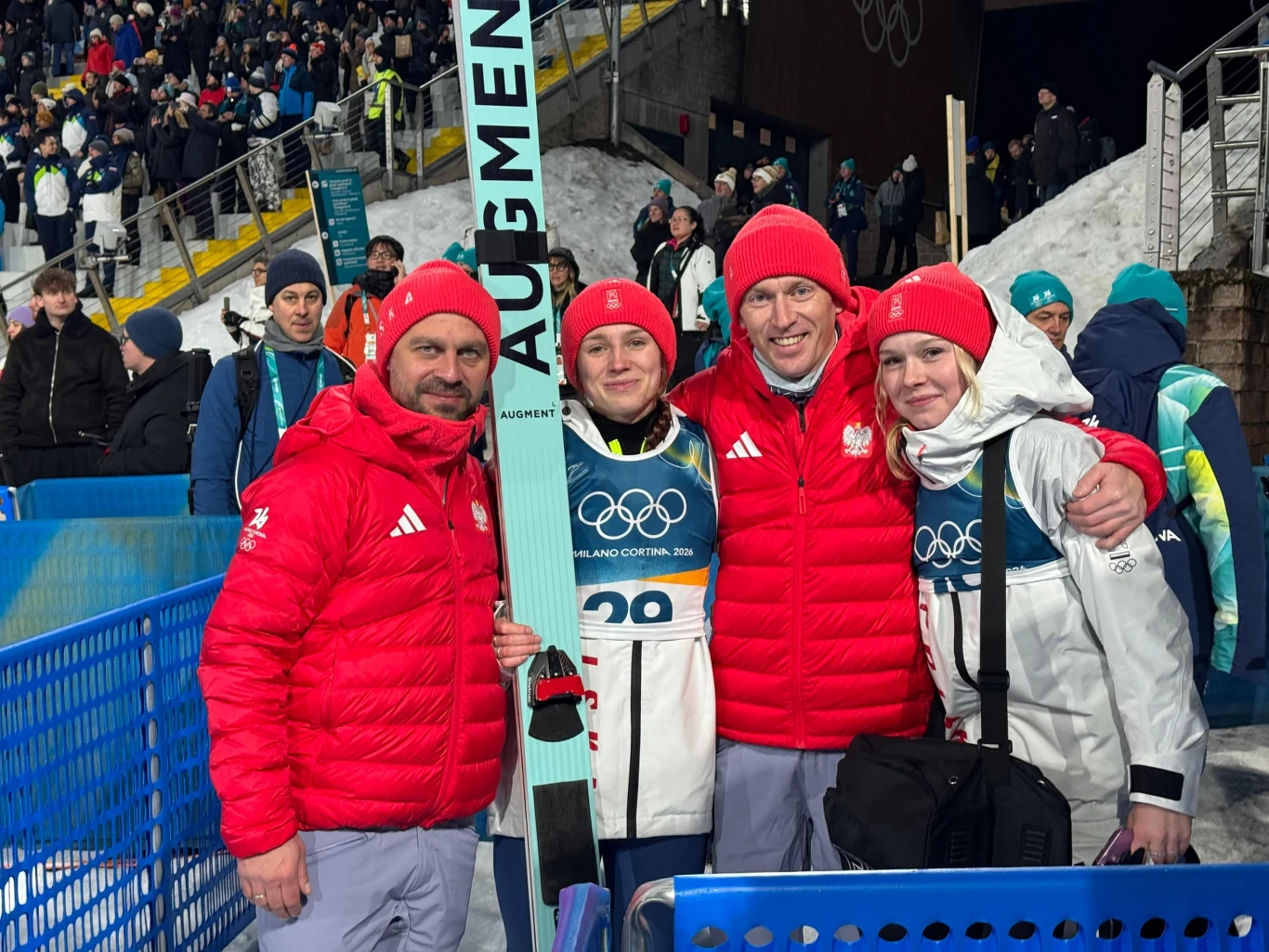 Zawodniczka drużyny skoczków narciarskich po konkursie zimowym ze sztabem, wszyscy ubrani w czerwone kurtki z polskimi emblematami, na tle olimpijskiego obiektu z widownią i śniegiem.