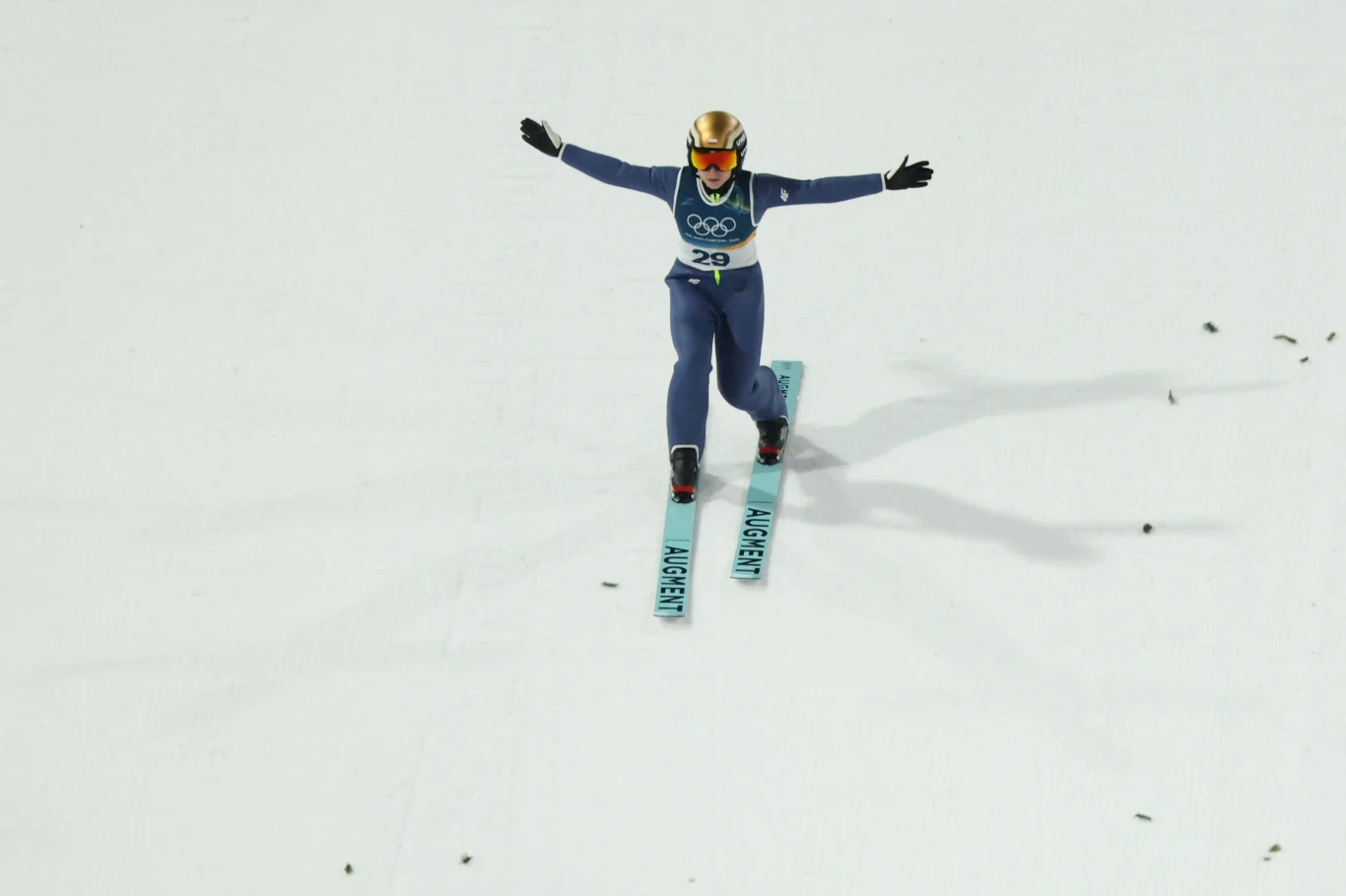 Skoczek narciarski w niebieskim kombinezonie i złotym kasku ląduje na śniegu z rozłożonymi ramionami, widoczne są narty z nazwą miejscowości, w tle białe podłoże skoczni.