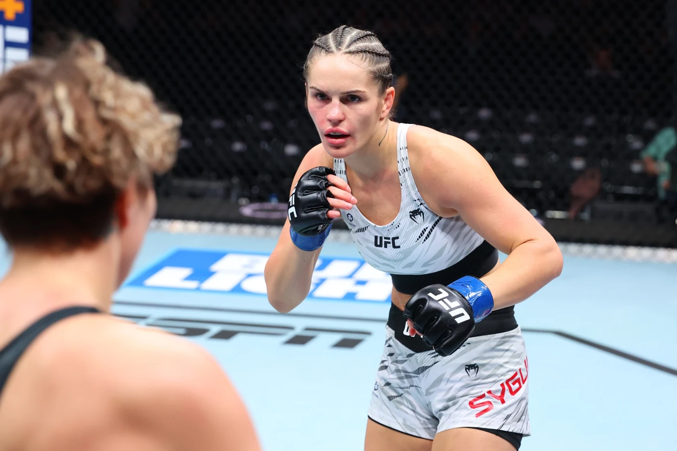 Zawodniczka UFC w sportowym stroju koncentruje się na walce z przeciwniczką w oktagonie. Na jej twarzy widać skupienie i determinację. W tle rozpoznawalna mata oraz ogrodzenie klatki ringowej.