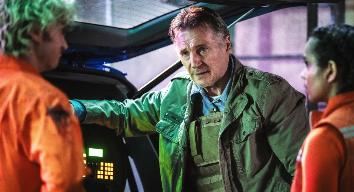 Joe Keery, Liam Neeson i Georgina Campbell w filmie "Skażenie" Trzech ludzi w rozmowie obok maszyny o wyglądzie technicznym, jedna osoba w kurtce i kamizelce taktycznej, pozostali w pomarańczowych kombinezonach roboczych.