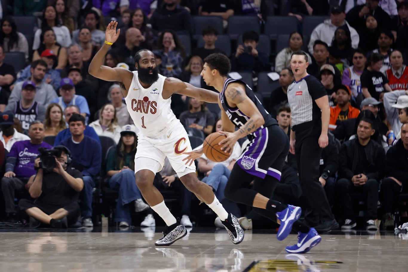 James Harden w barwach Cleveland Cavaliers Dwóch koszykarzy podczas meczu NBA, zawodnik w białym stroju drużyny Cavaliers stara się zatrzymać zawodnika w czarnym stroju z piłką z drużyny Sacramento Kings, w tle widownia i sędzia na parkiecie.