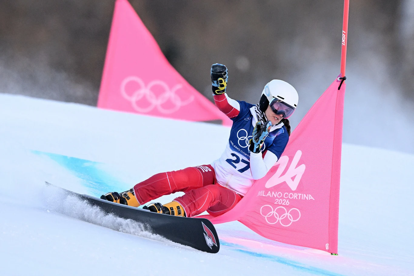 Aleksandra Król-Walas Zawodnik na snowboardzie w trakcie dynamicznego skrętu na stoku, ubrany w biały kask, gogle oraz kombinezon startowy z numerem 27, przejeżdża obok różowego pachołka z logo igrzysk olimpijskich Milano Cortina 2026.