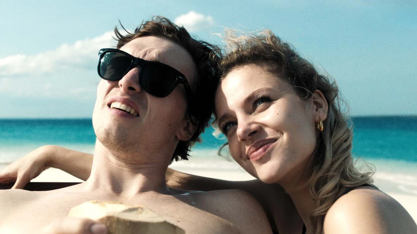 Tomasz Włosok i Vanessa Aleksander w filmie "Król dopalaczy" Para młodych ludzi relaksuje się na plaży przy lazurowym morzu. Mężczyzna w okularach przeciwsłonecznych i kobieta z uśmiechem na twarzy siedzą blisko siebie, ciesząc się słoneczną pogodą i letnią atmosferą.