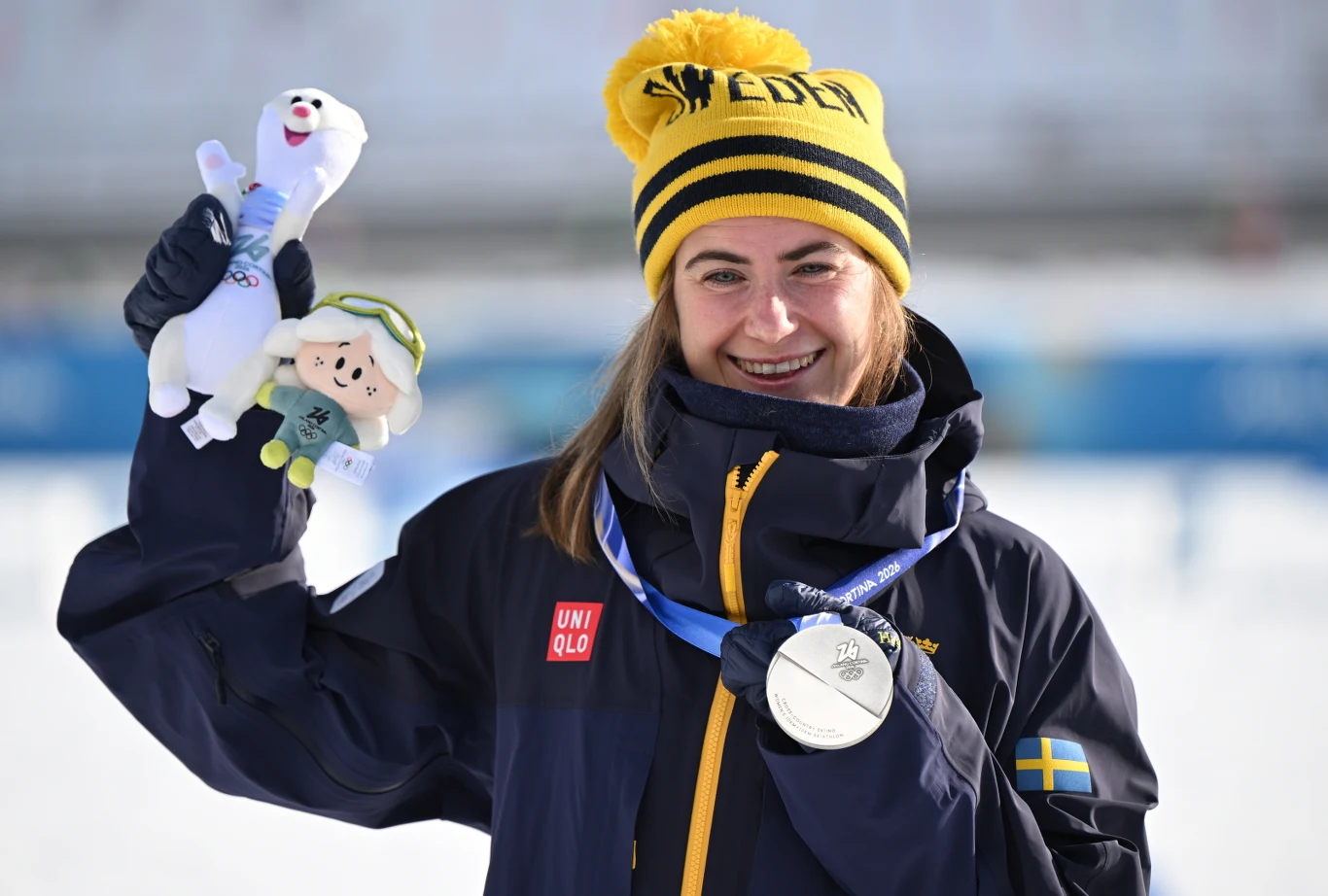 Ebba Andersson wywalczyła srebro w skiathlonie Zadowolona zawodniczka w czapce z napisem Sweden prezentuje srebrny medal oraz maskotki, stojąc na tle zimowej scenerii. Na kurtce widoczna flaga Szwecji, kobieta uśmiecha się szeroko.