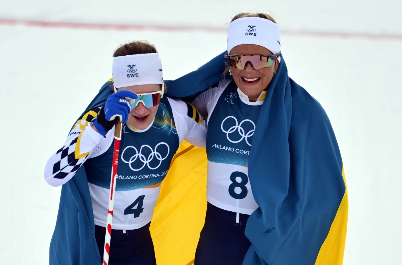 Szwedki zdominowały skiathlon Dwie zawodniczki narciarstwa biegowego ubranej w stroje sportowe i opaski z napisem SWE, owinięte w niebiesko-żółte flagi, uśmiechnięte po zakończonym biegu olimpijskim.